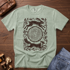 A sage green crewneck t-shirt with a dark brown graphic print of a hare, fox, and tree stump, styled in a flat lay on a wooden surface with a brown leather bag, mushrooms, and blue jeans.